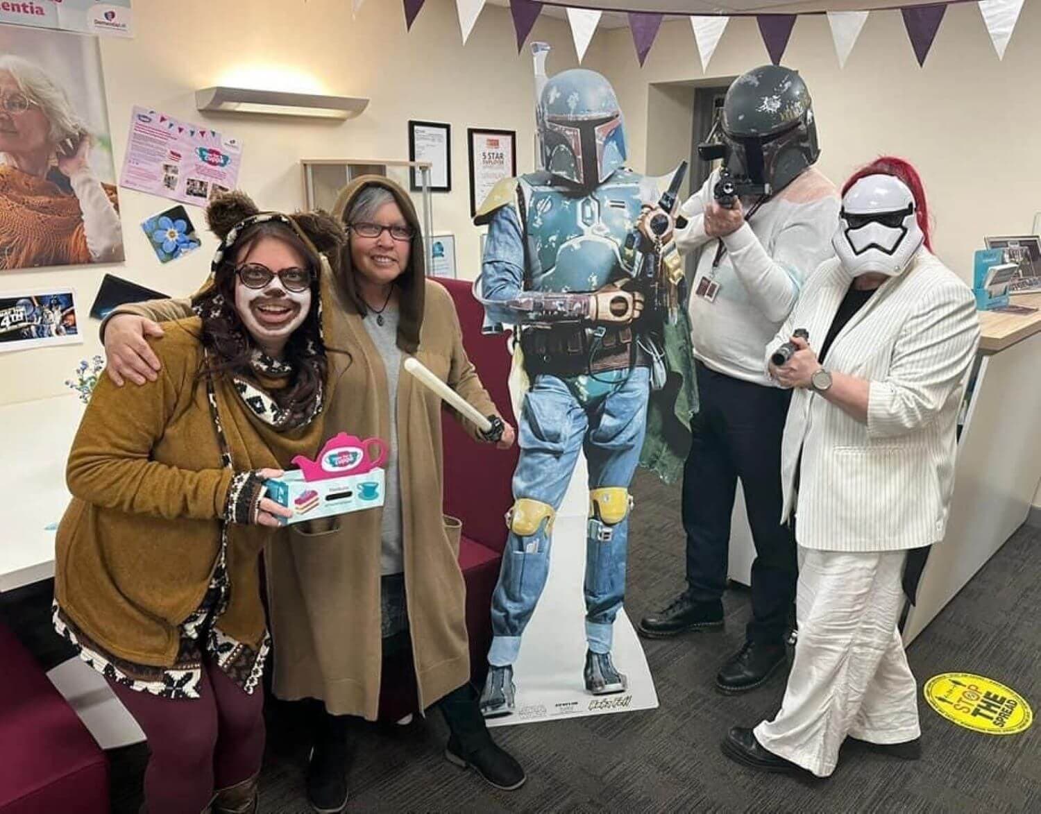 Four people in costumes, including Star Wars characters, pose around a cardboard standee in an office decorated for Halloween. - Home Instead