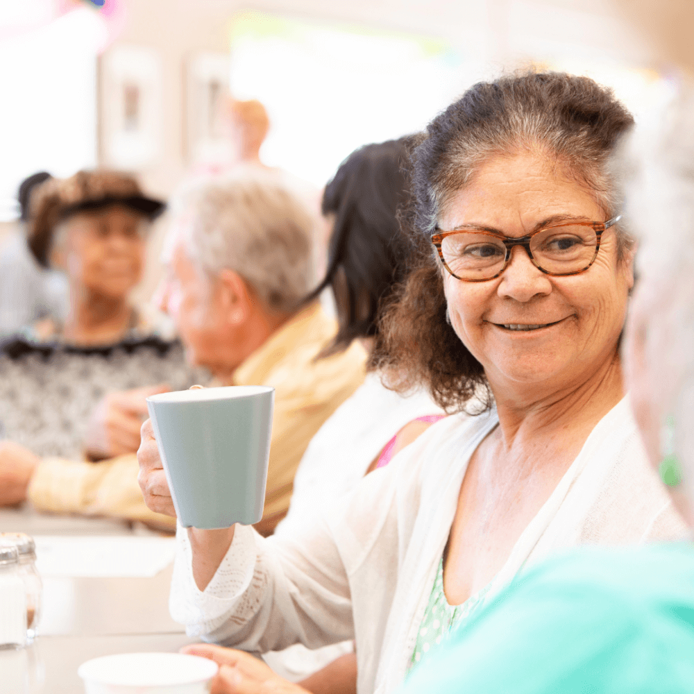 Smiling woman with glasses holding a cup, sitting with a group of seniors at a social gathering. - Home Instead