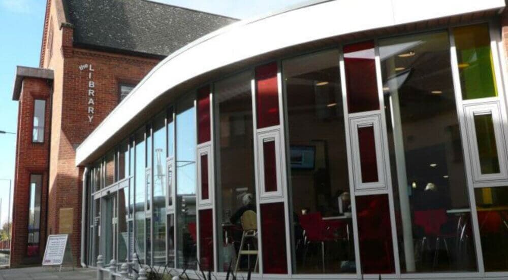 Exterior of a modern red-brick library with large curved glass windows and colorful panels, showing the word "LIBRARY. - Home Instead
