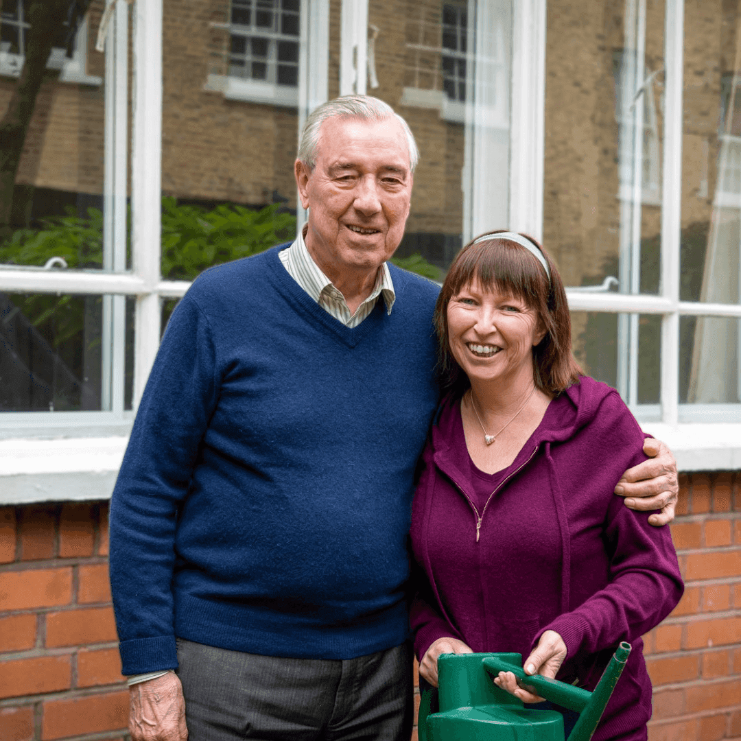 An elderly man and a middle-aged woman standing and smiling with a watering can outside a building. - Home Instead