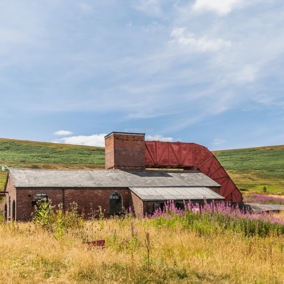Old brick building with a large rusted structure attached, set in a field with hills and a partly cloudy sky in the background. - Home Instead