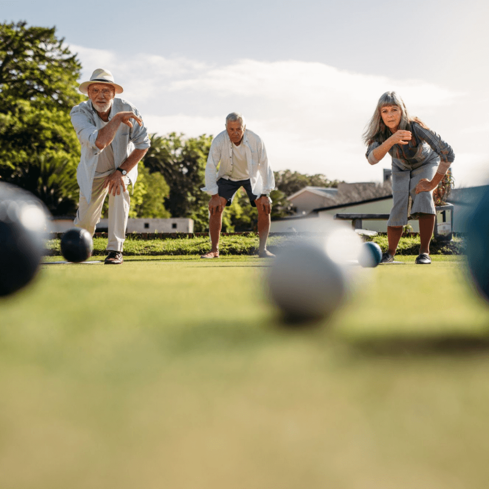 Three elderly people playing bocce ball on a sunny day, focusing and preparing to throw their balls on the lawn. - Home Instead