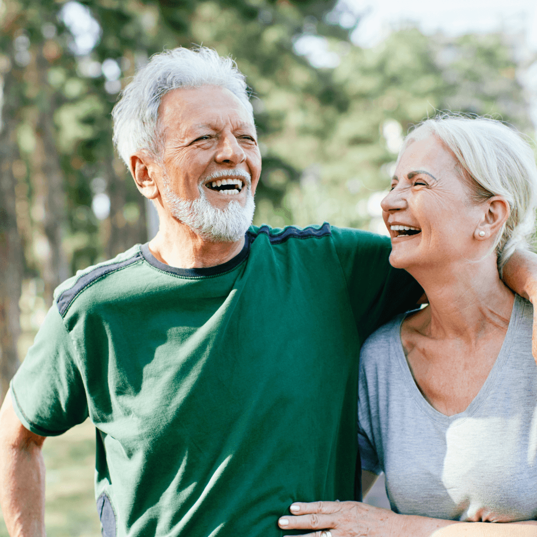 An elderly couple, smiling and laughing together outdoors, with trees in the background. - Home Instead