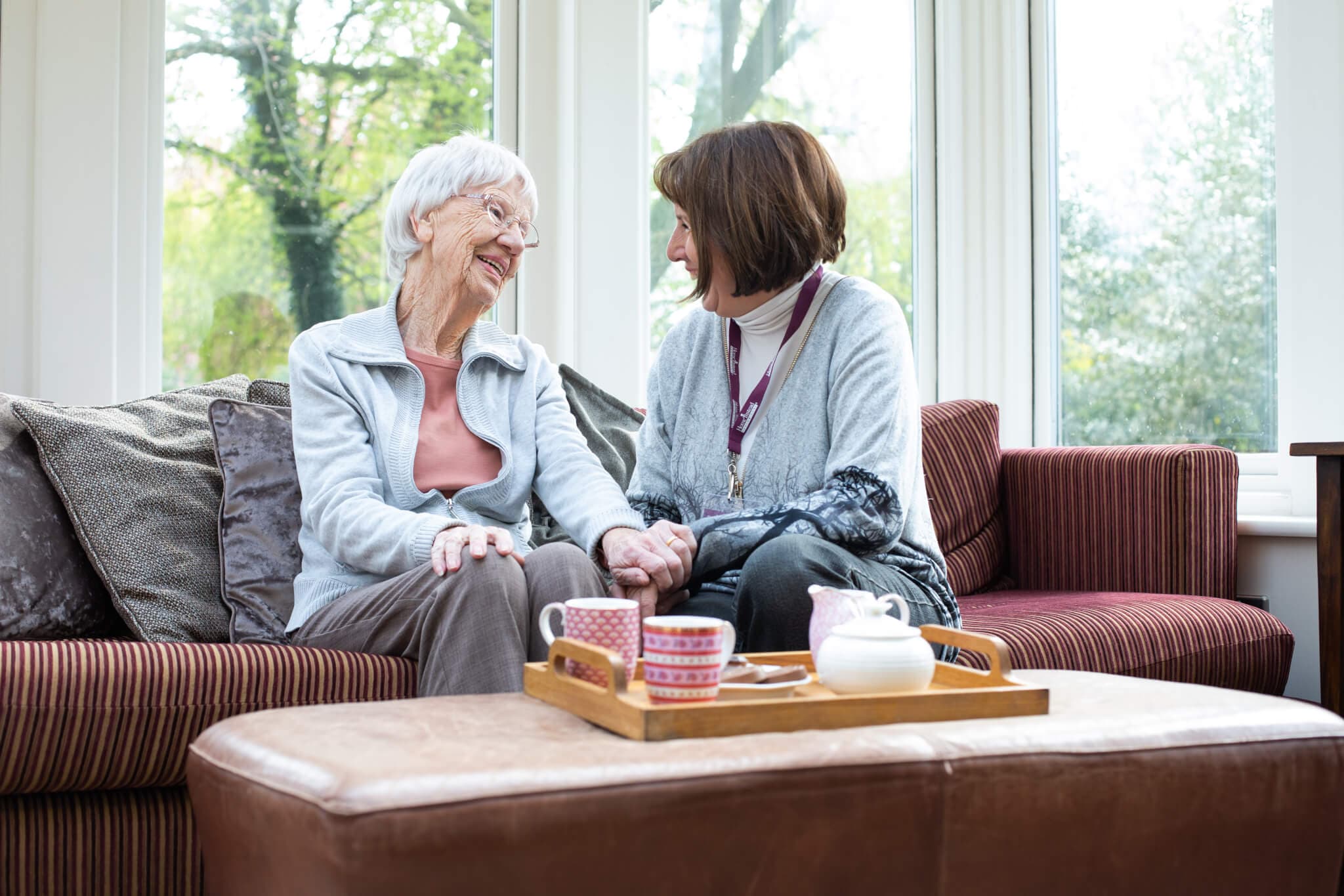 Two women sitting on a couch, smiling and holding hands, with tea on a tray on the table in front of them. - Home Instead