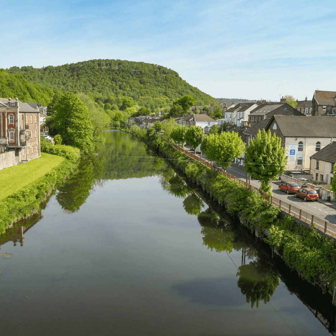 A calm river reflecting green hills and houses on both sides under a clear blue sky. - Home Instead