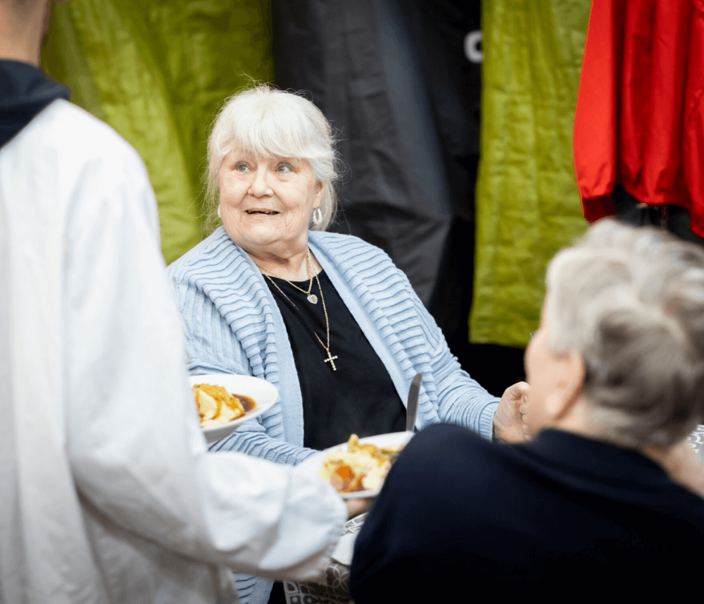 An elderly woman with white hair, wearing a blue sweater, smiles while seated at a table with food in front of her. - Home Instead