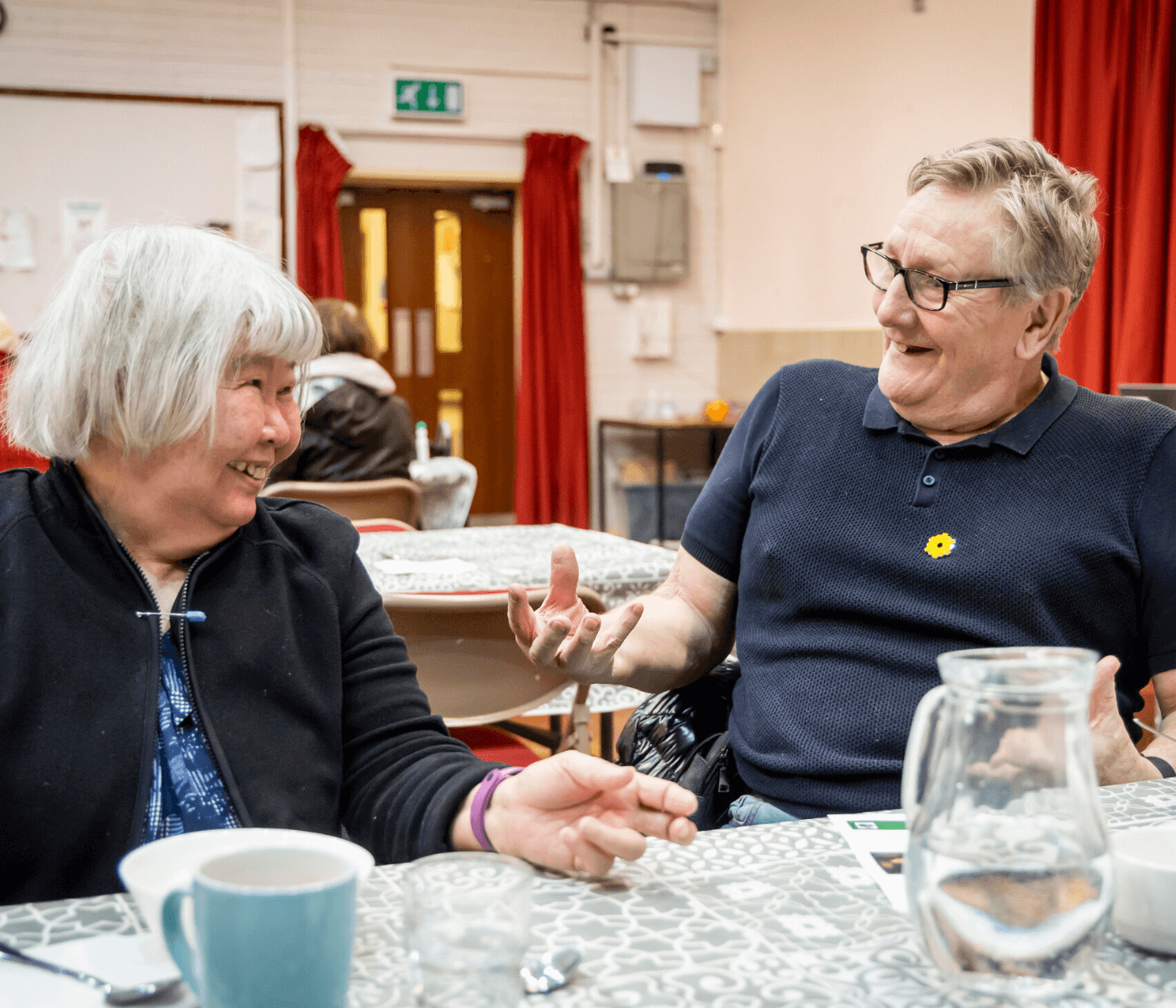 Two older adults are laughing and chatting at a table with a patterned tablecloth in a casual indoor setting. - Home Instead
