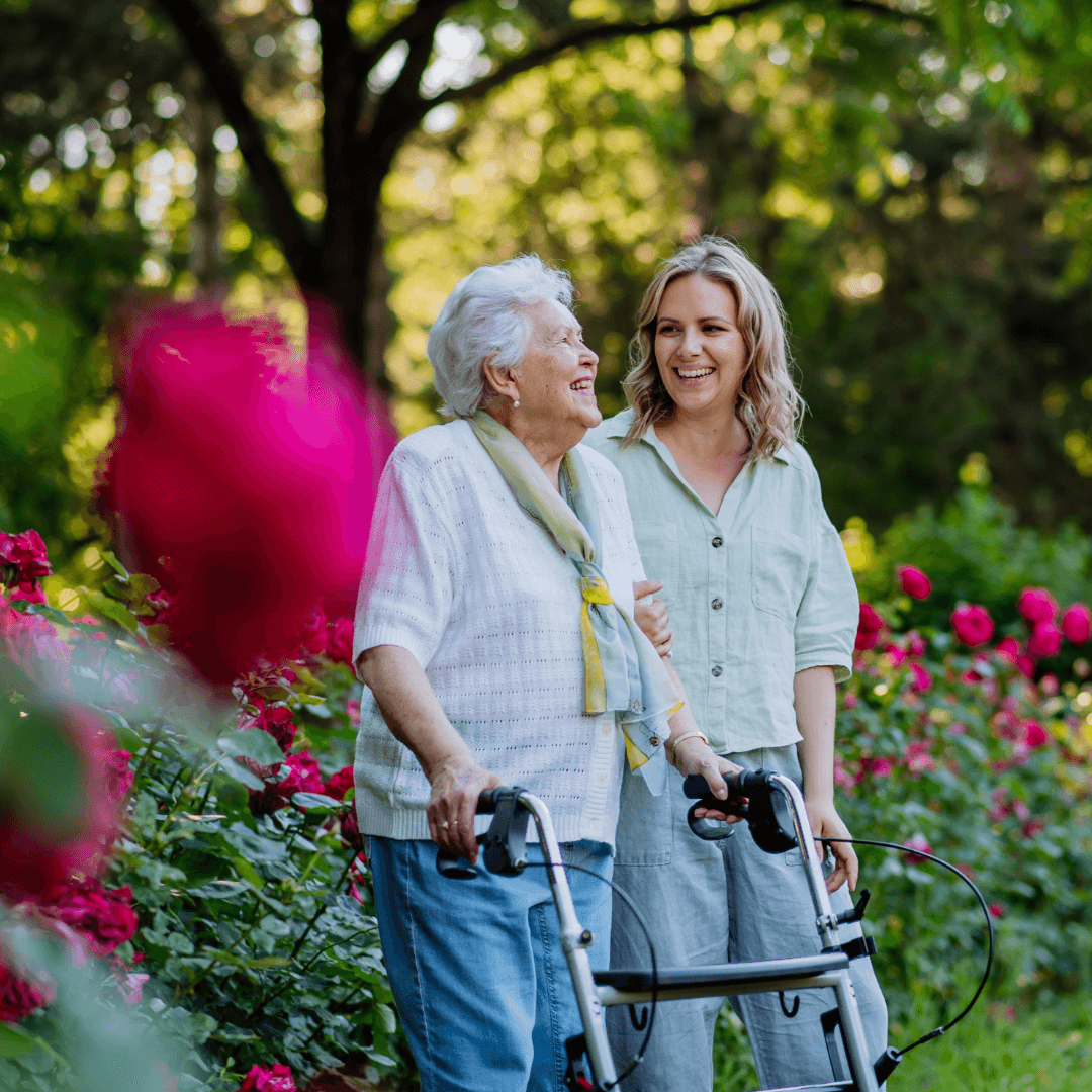 Elderly woman with a walker and a young woman smiling together in a garden with vibrant flowers. - Home Instead