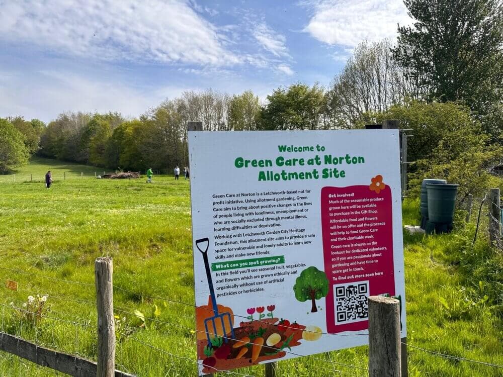 A sign at Green Care at Norton Allotment Site, with a field and people working in the background on a sunny day. - Home Instead