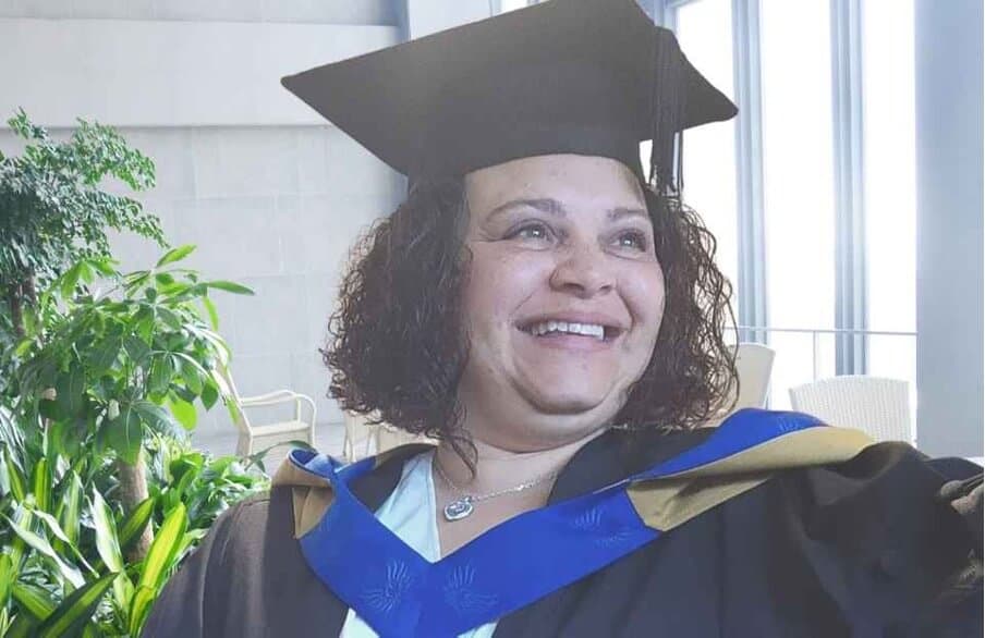 A person wearing a graduation cap and gown, smiling and looking to the side. Green plants are in the background. - Home Instead