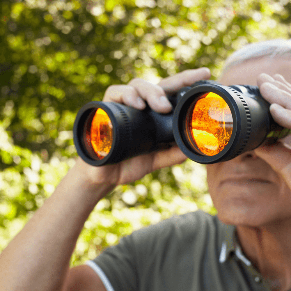 Elderly man looking through binoculars with orange-tinted lenses in a forested area. - Home Instead