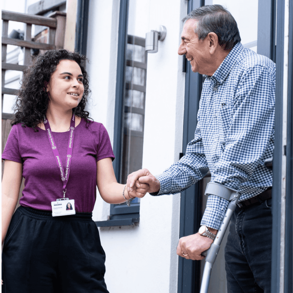 A young woman with a badge shakes hands with an older man using crutches, both smiling outside a building. - Home Instead