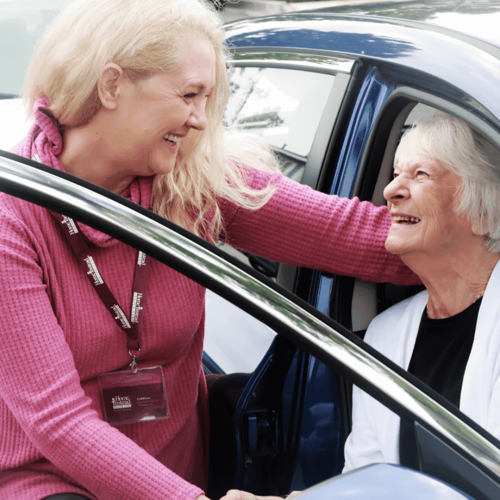 A caregiver helps an elderly woman out of a car, both smiling warmly at each other. - Home Instead