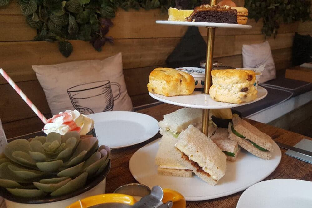 A three-tiered stand with scones, pastries, and sandwiches on a wooden table next to a plant and a cupcake. - Home Instead