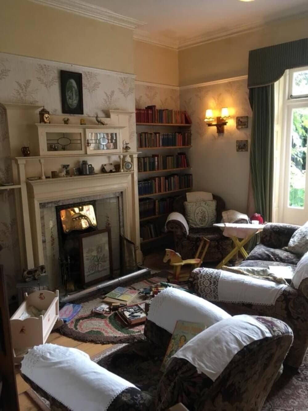 Cozy living room with vintage furniture, fireplace, bookshelves, scattered toys, and a large window. - Home Instead