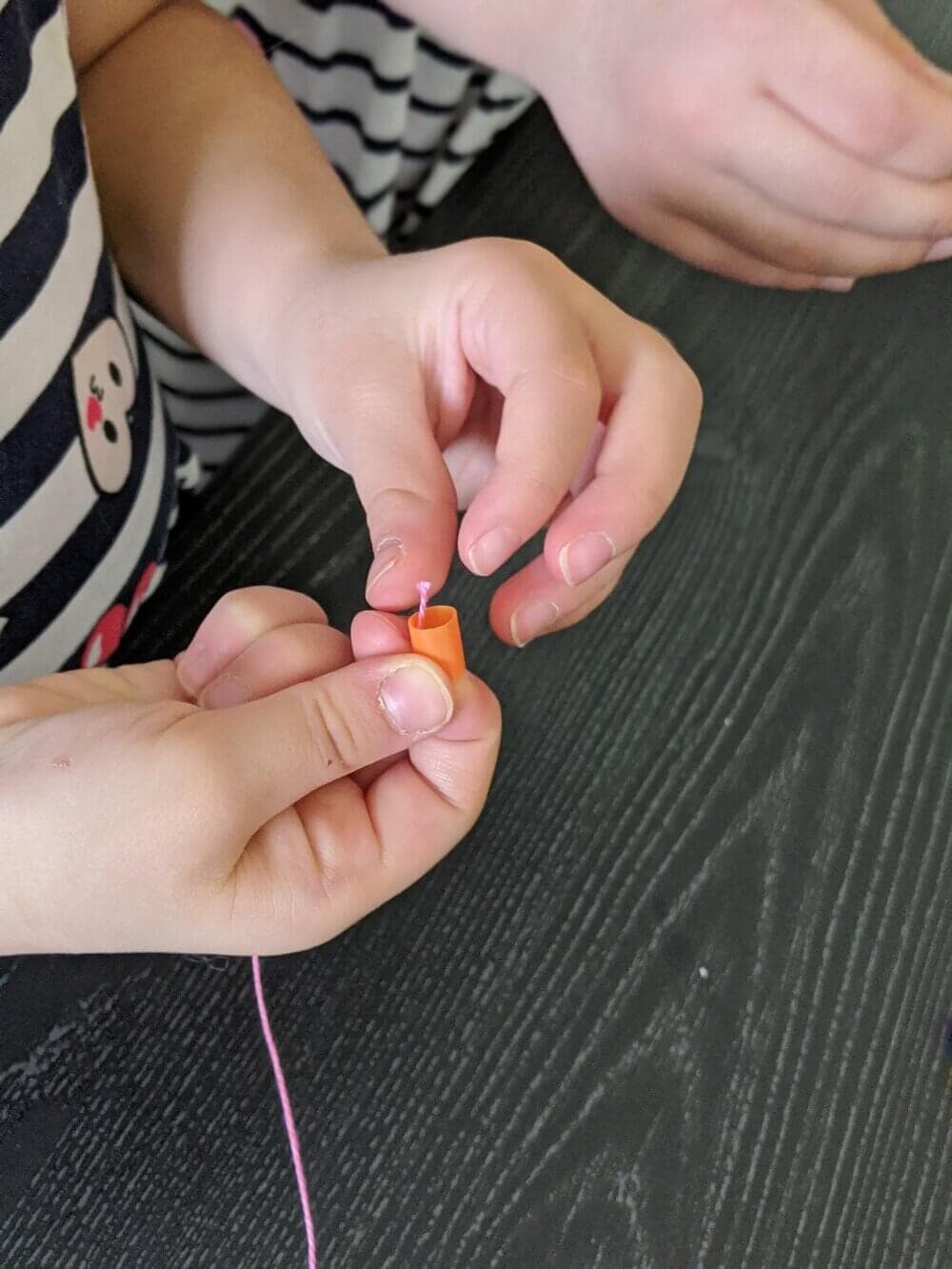 Child threading a needle with pink thread, wearing a striped shirt, against a dark table background. - Home Instead