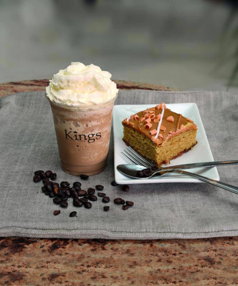 Iced coffee topped with whipped cream in a cup labeled "Kings" beside a cake slice on a plate with a fork and spoon. - Home Instead