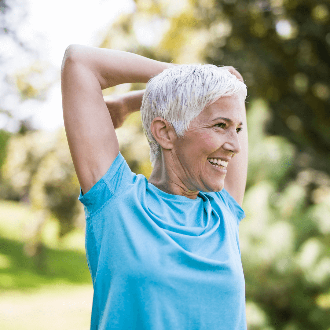 Elderly woman with short gray hair stretching her arms and smiling outdoors in a park, wearing a blue shirt. - Home Instead