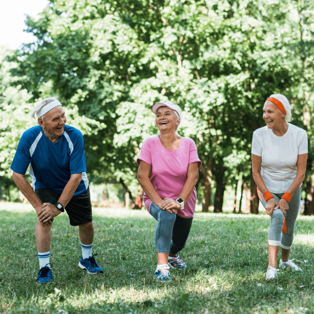 Three elderly people in sportswear laughing and stretching together outdoors in a park. - Home Instead