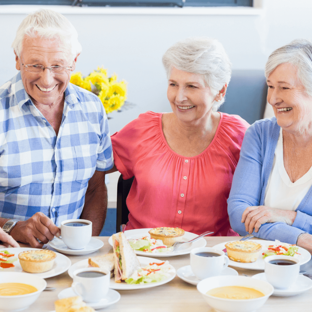 Three older adults enjoying coffee and pastries while smiling and chatting at a table. - Home Instead