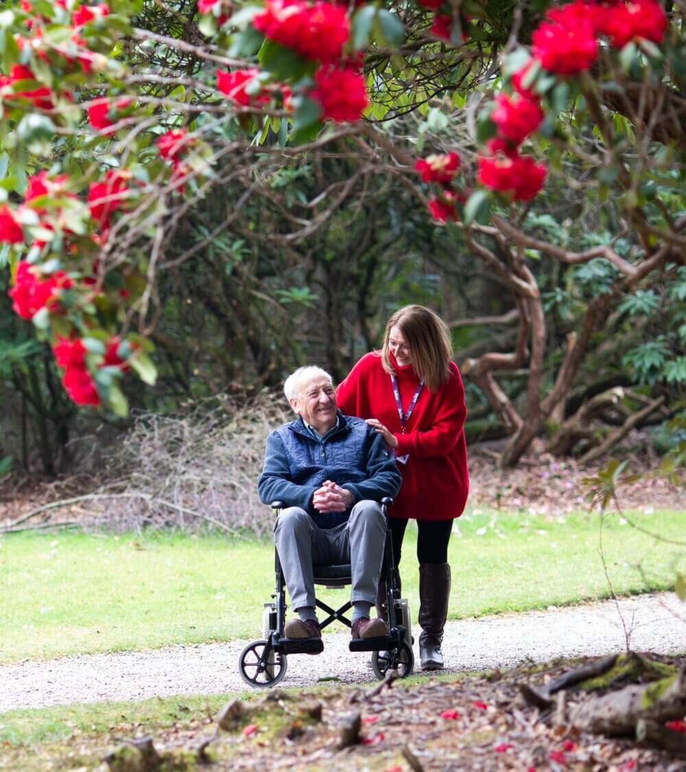 A woman assists a smiling older man in a wheelchair as they enjoy a walk in a park with vibrant red flowers. - Home Instead