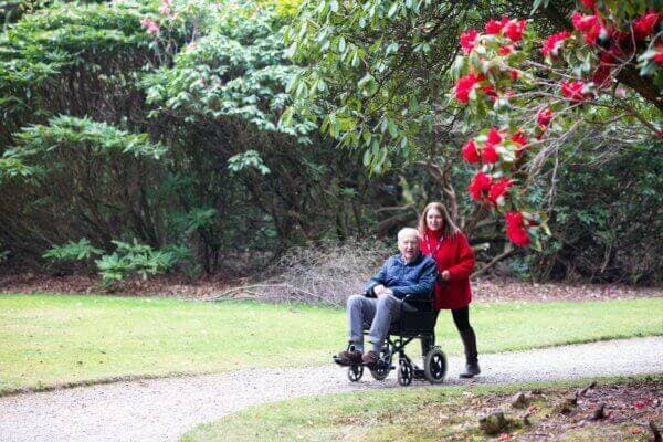 A woman in a red coat pushes an elderly man in a wheelchair along a park path with greenery and red flowers around them. - Home Instead