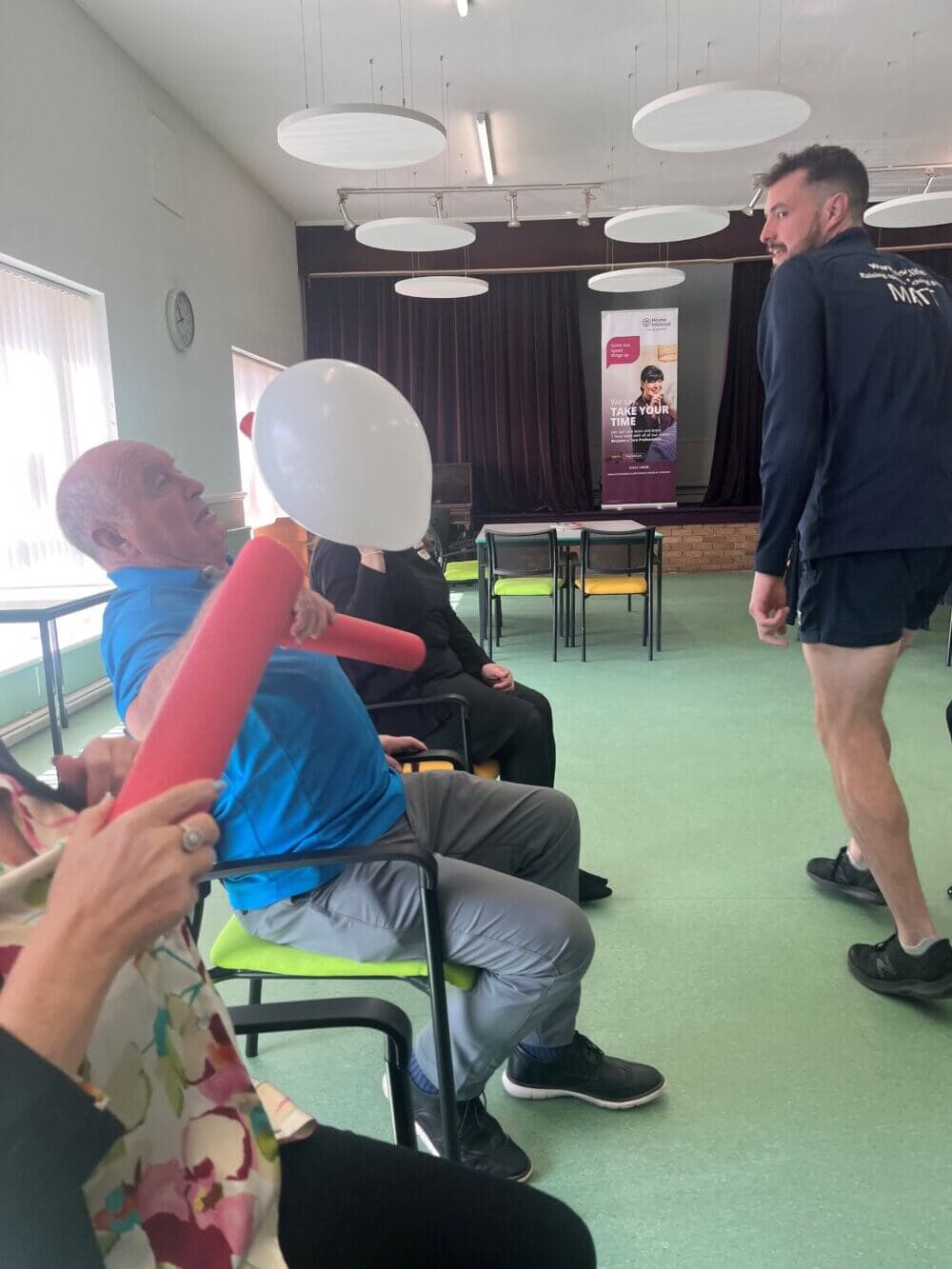 People engaging in a fun balloon activity in a room with large windows and a person standing in the background. - Home Instead