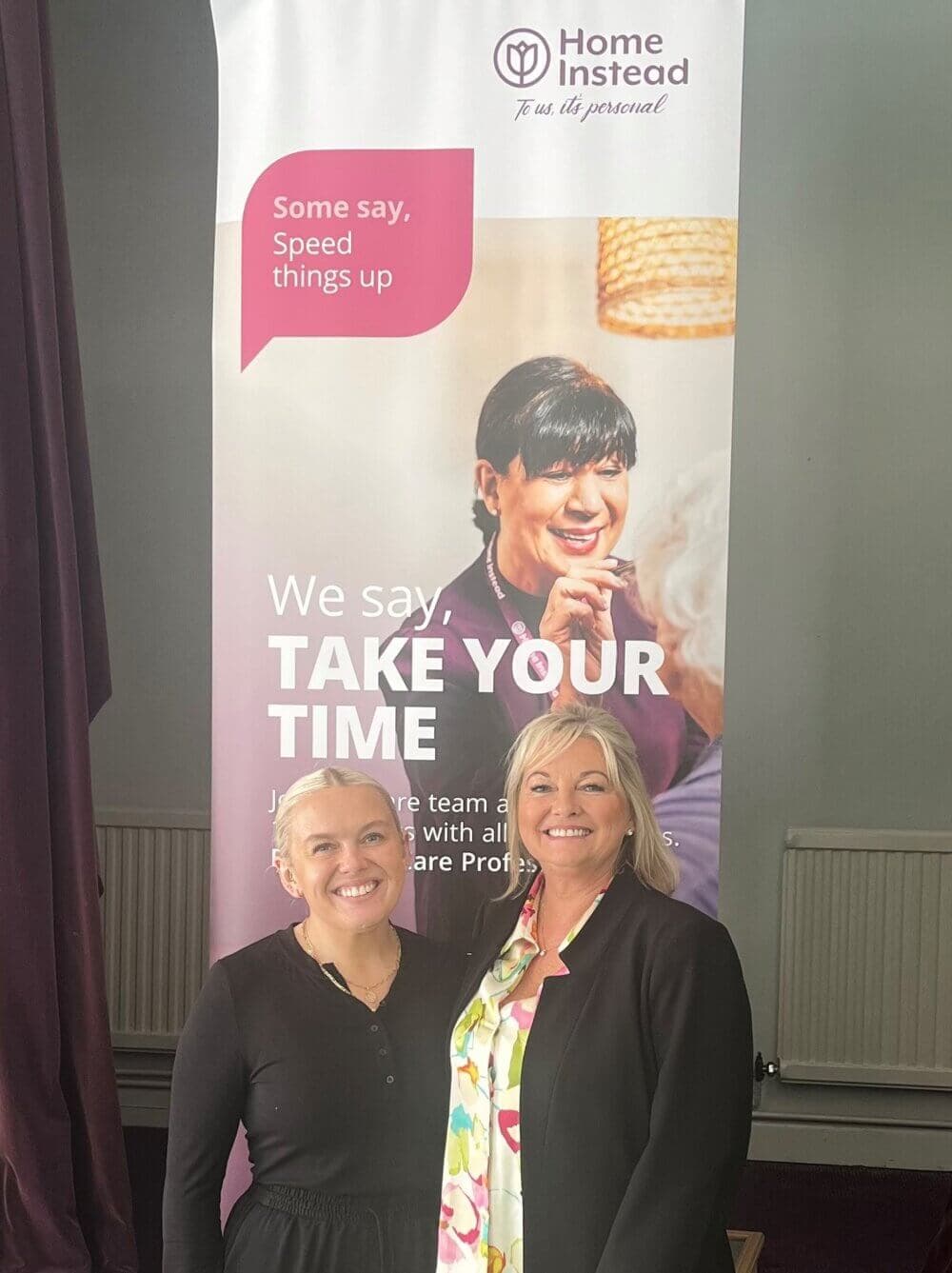 Two women smiling in front of a Home Instead banner that says "We say, TAKE YOUR TIME. - Home Instead