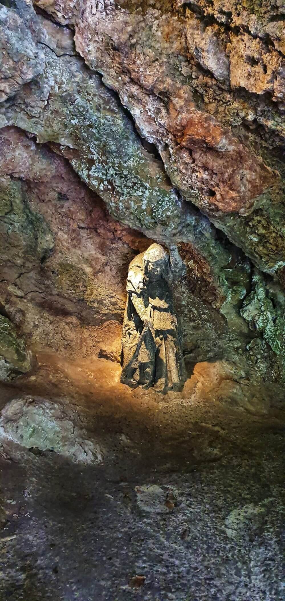 Stone statue of a cloaked figure sitting and holding its head in a rocky cave, illuminated by light. - Home Instead