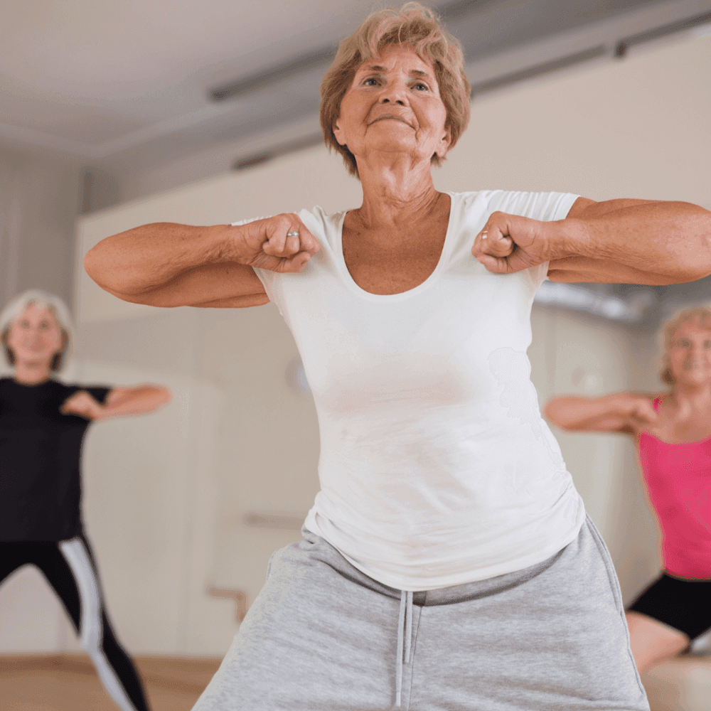 Elderly women in workout class, with the focus on one woman in a white shirt lifting weights, the others in the background. - Home Instead