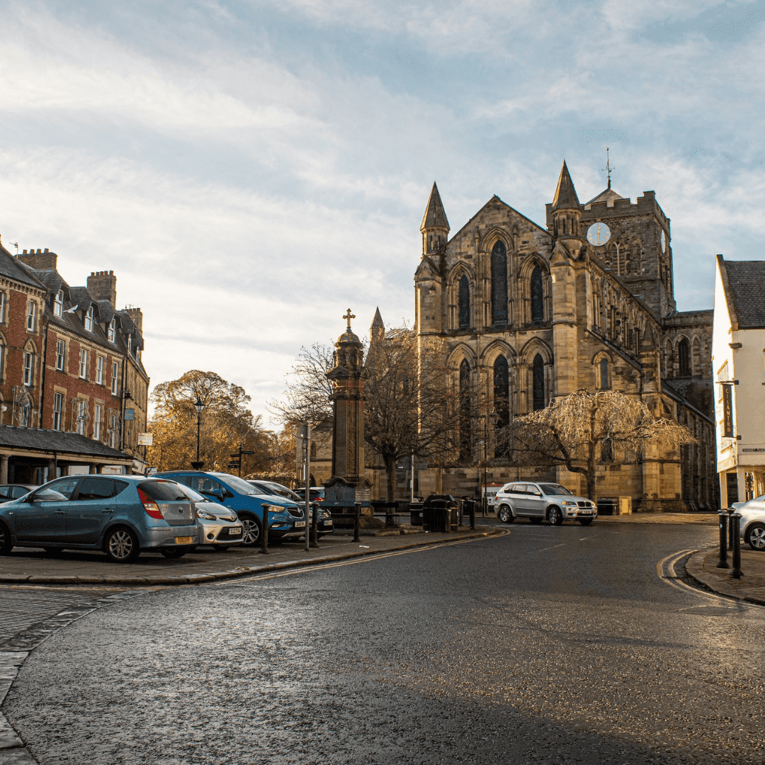Historic stone church with pointed arches and a clock tower, viewed from a street lined with parked cars and old buildings. - Home Instead