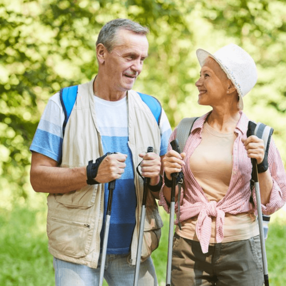 Two smiling elderly adults with backpacks and hiking poles enjoying a walk in a green outdoor setting. - Home Instead