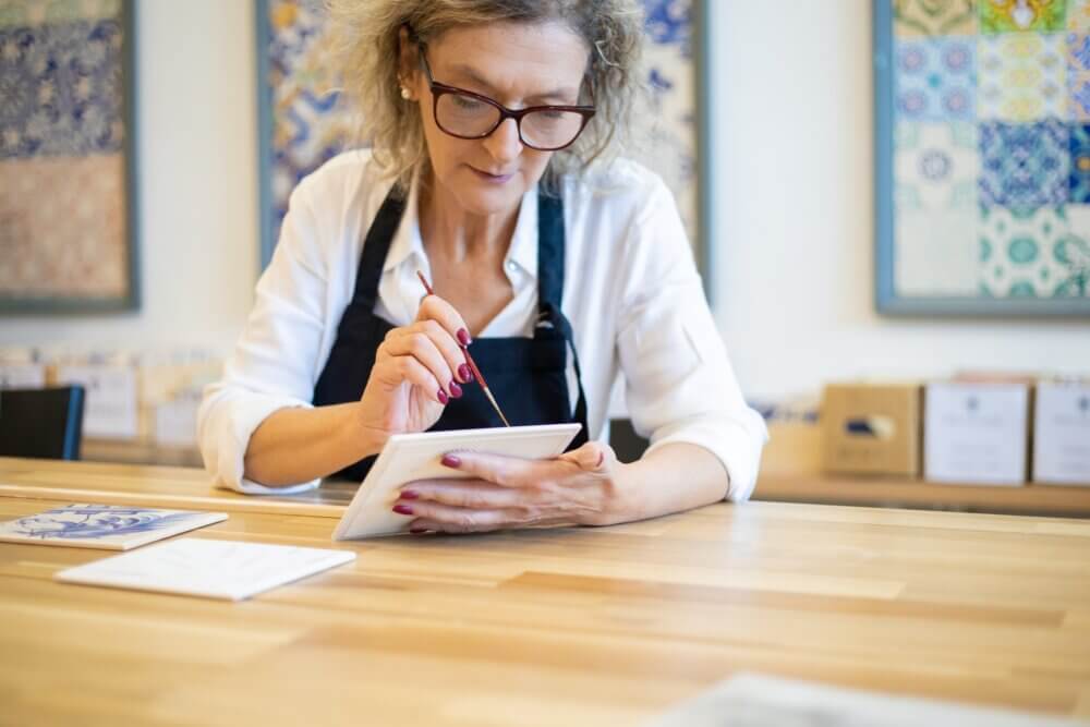 A woman in glasses and an apron paints a tile while sitting at a wooden table in a bright room with colorful decor. - Home Instead
