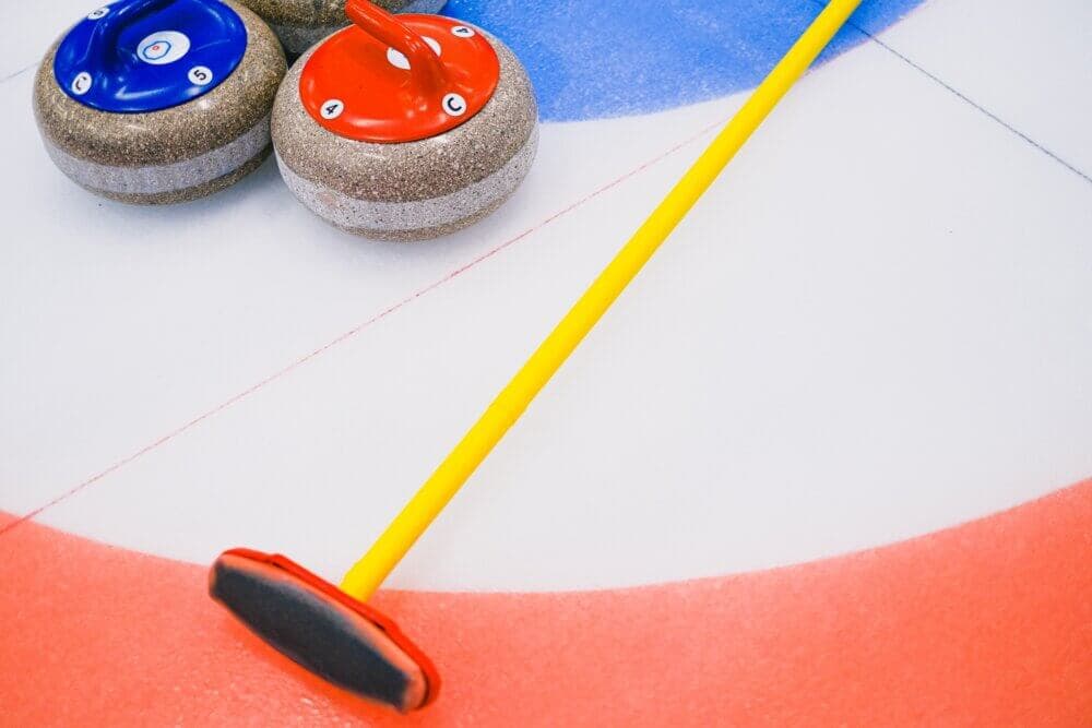 Curling stones and a broom on an ice rink, set in the target area with red, blue, and white rings. - Home Instead