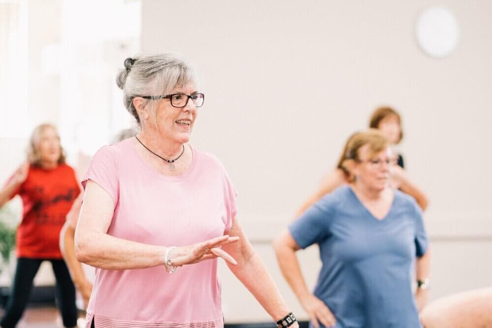Elderly women participating in a group exercise class, one in a pink shirt and glasses in the foreground. - Home Instead