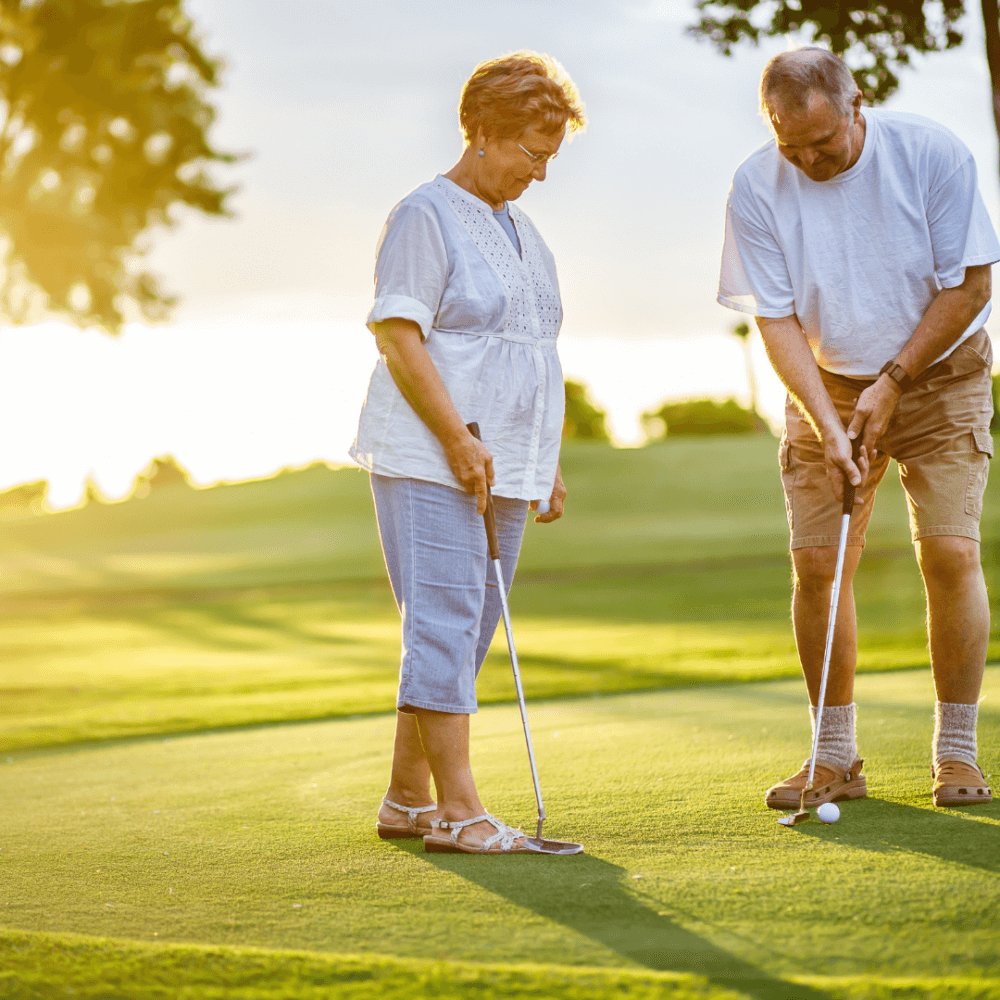 An older couple playing golf on a sunny day. The woman is preparing to putt while the man watches and offers advice. - Home Instead