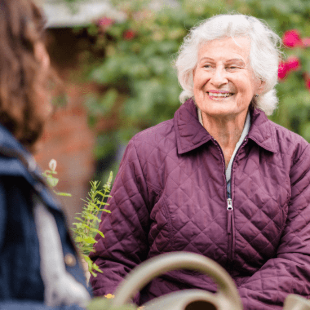 Elderly woman in purple jacket smiling while sitting and talking with another person outdoors in a garden. - Home Instead