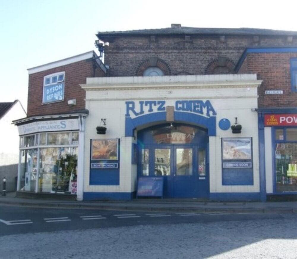 Front view of the Ritz Cinema building with posters on display, flanked by two small shops on either side. - Home Instead