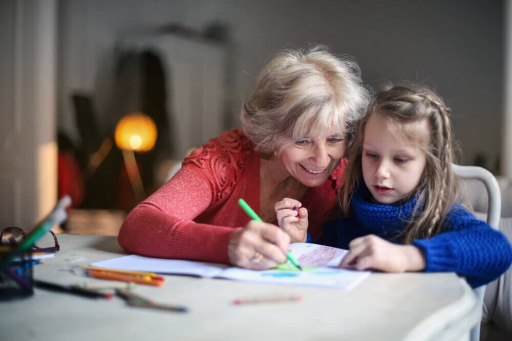 An elderly woman and a young girl are sitting at a table, drawing together with colored pencils. - Home Instead