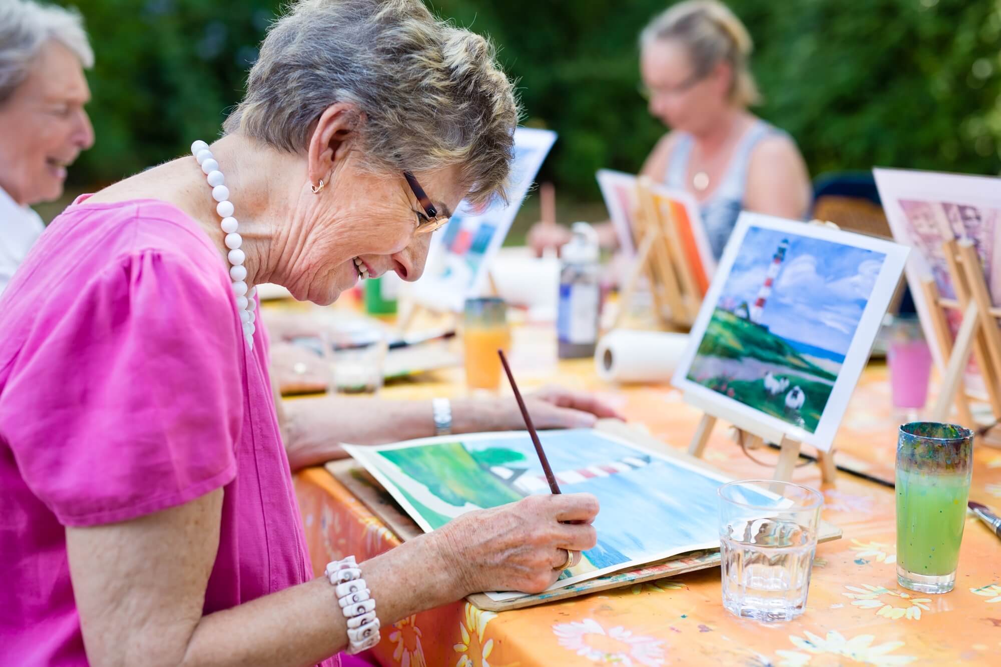 Elderly woman smiling and painting outdoors with others, colorful paintings on easels in the background. - Home Instead