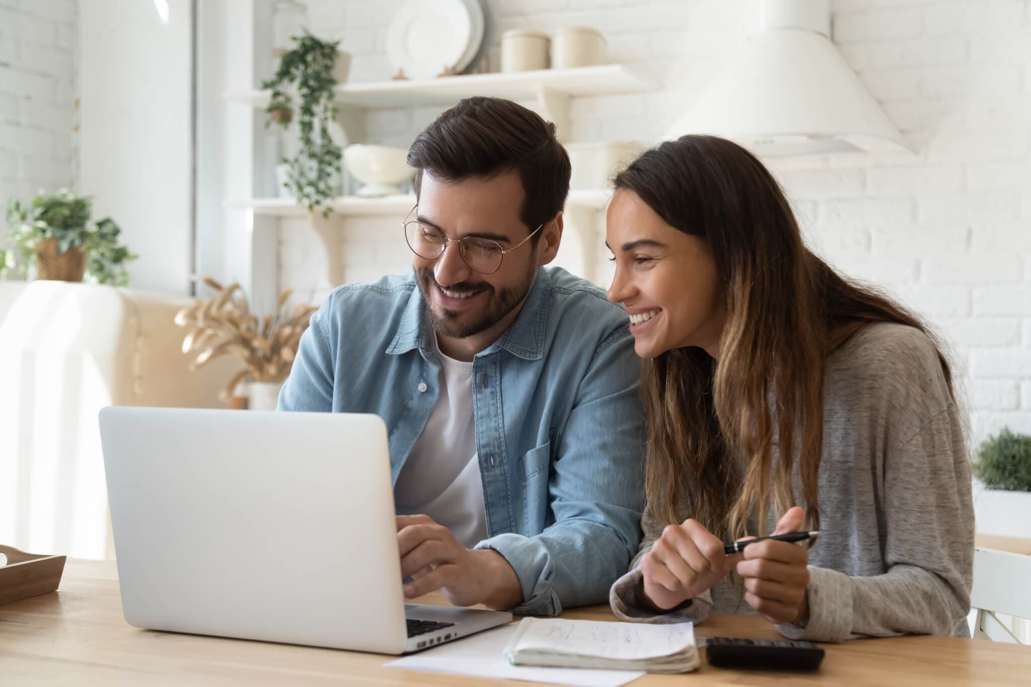 A smiling couple sits at a table, looking at a laptop. They are in a bright room with plants and kitchen items in the background. - Home Instead
