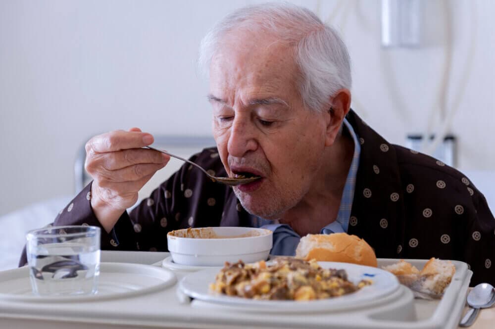 Elderly man eats soup in a hospital, with a tray of food and a glass of water in front of him. - Home Instead