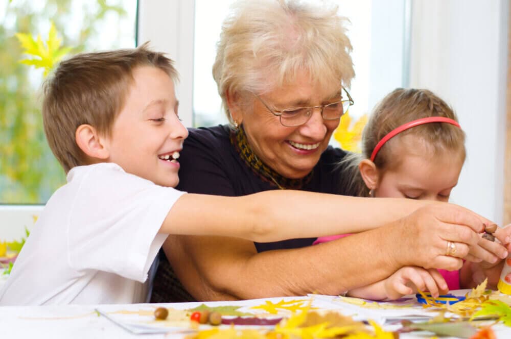 An elderly woman smiles and helps a young boy and girl with an art project involving leaves and various craft materials. - Home Instead