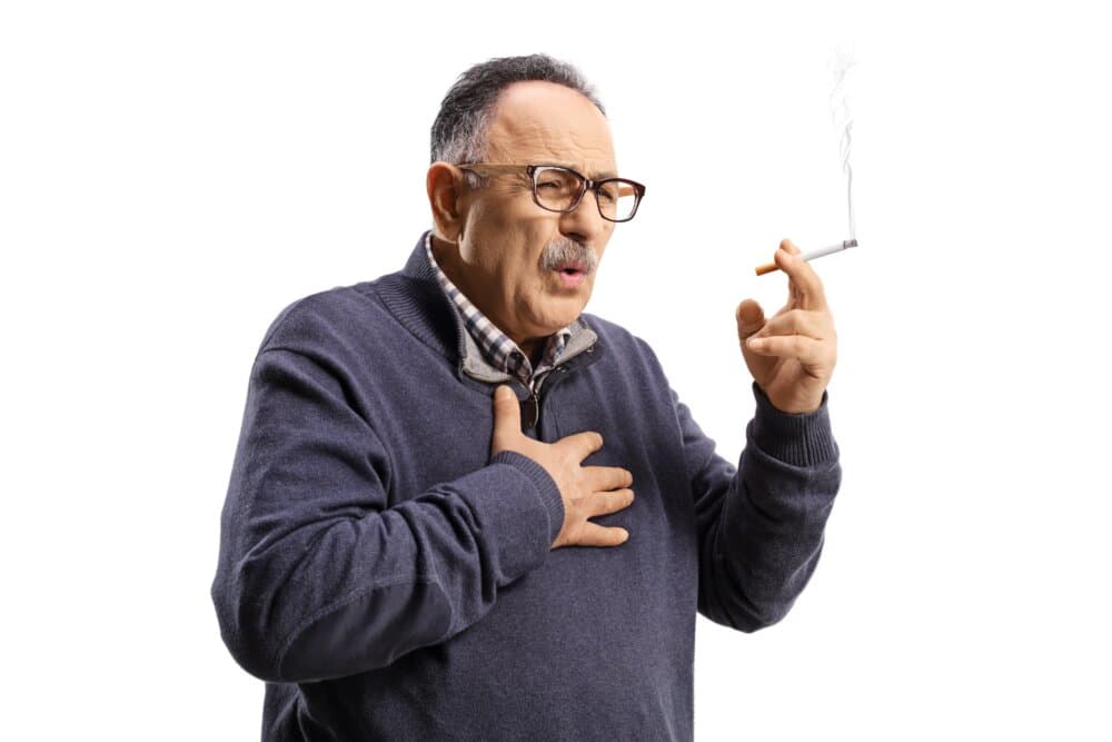 An older man with glasses holds his chest with one hand and a lit cigarette with the other, appearing to cough. - Home Instead
