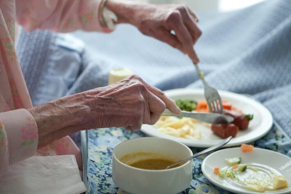 An elderly person in pink pajamas eating a meal with vegetables and a bowl of soup, on a table with a blue floral tablecloth. - Home Instead