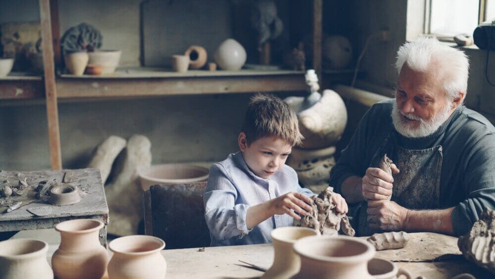 A child and an elderly man work with clay in a pottery workshop surrounded by finished pots and tools. - Home Instead