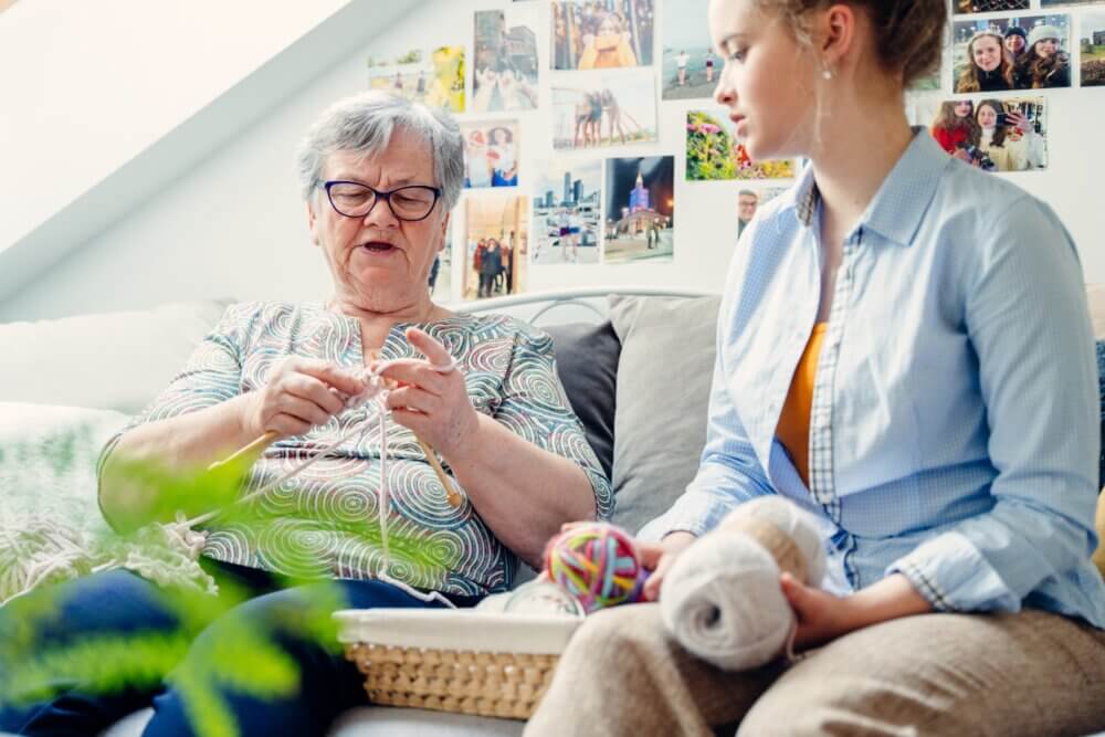 Elderly woman knitting while younger woman watches, seated on a couch with photo-covered wall in the background. - Home Instead