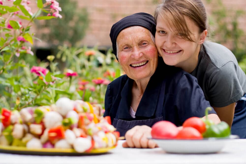 A smiling elderly woman and a young woman pose outdoors near a table with vegetables and flower plants. - Home Instead