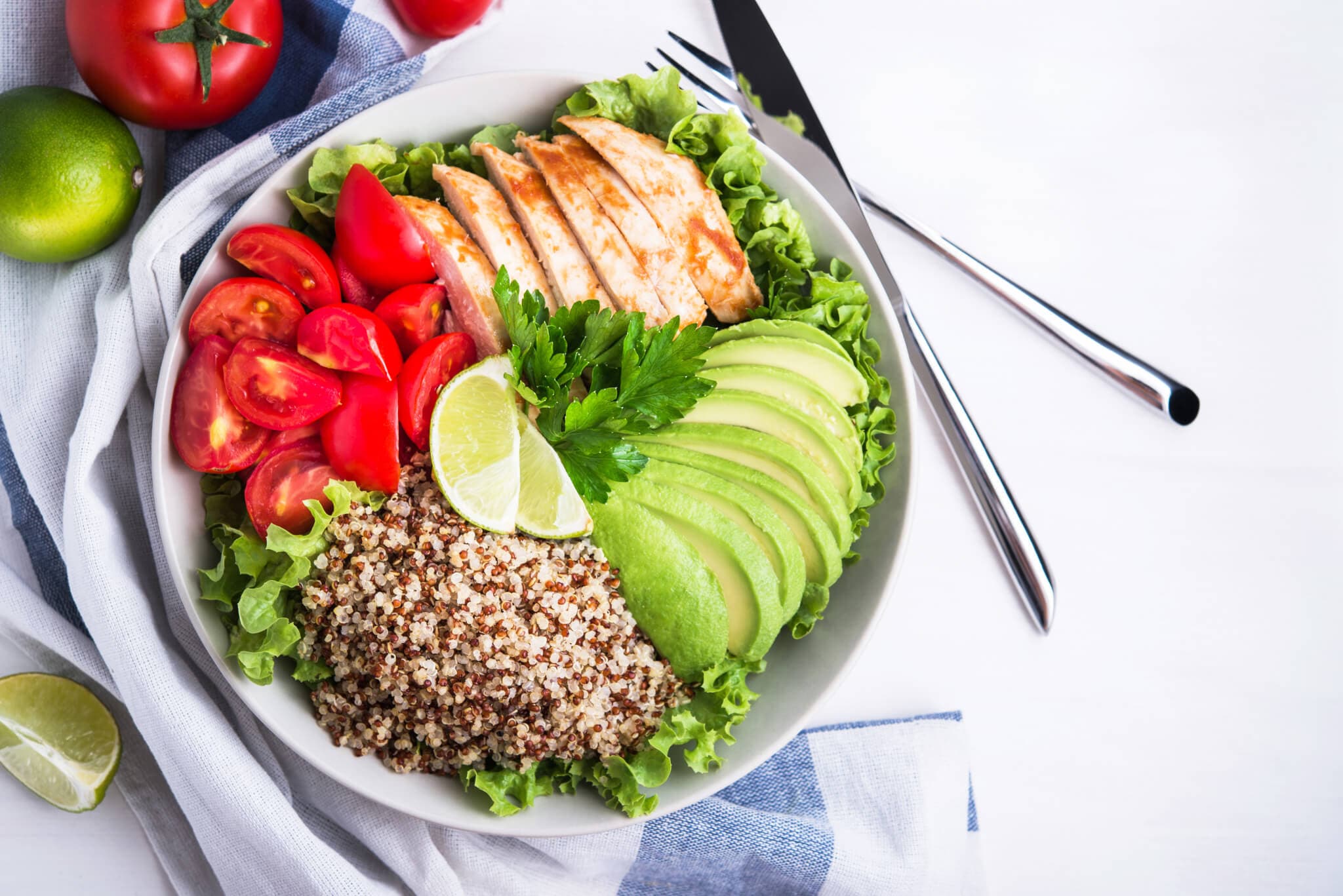 A bowl with sliced chicken, avocado, tomato, quinoa, lime wedges, and greens on a striped cloth with utensils beside it. - Home Instead