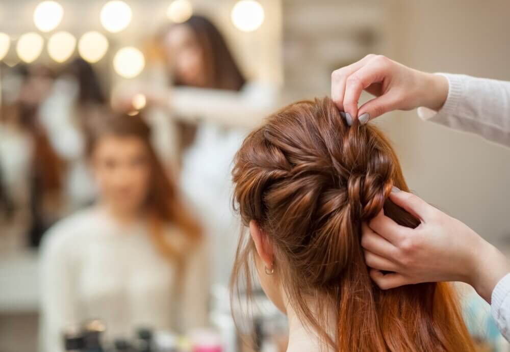 A hairstylist braiding a woman's hair in a salon, with the woman reflected in a mirror in the background. - Home Instead